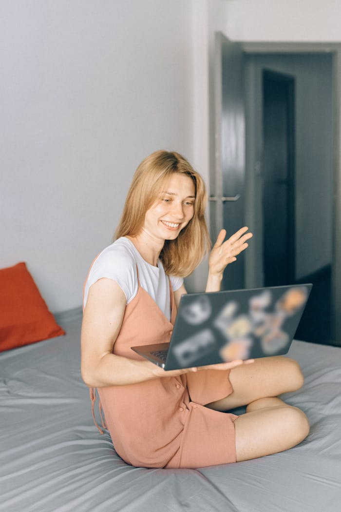 Woman sitting on bed working on laptop, smiling during a video call in cozy home setting.